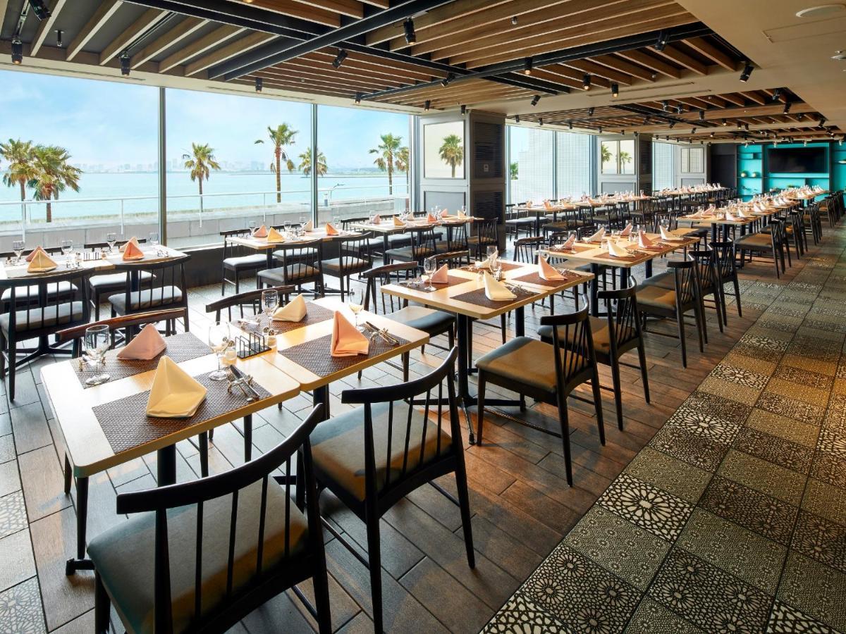 Hotel Grand Nikko Tokyo Bay Maihama Urayasu Exterior foto The photo shows a modern restaurant interior featuring rows of neatly arranged tables and chairs. The large windows provide a scenic view of palm trees and what appears to be a body of water in the background, suggesting a coastal location. Each tabl