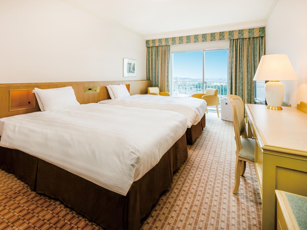 Hotel Grand Nikko Tokyo Bay Maihama Urayasu Exterior foto The photo shows a hotel room featuring two twin beds with white linens and dark brown bed skirts. The room has a light, neutral-colored wall and carpet. There is a window with curtains that let in natural light, offering a view outside. A small desk 